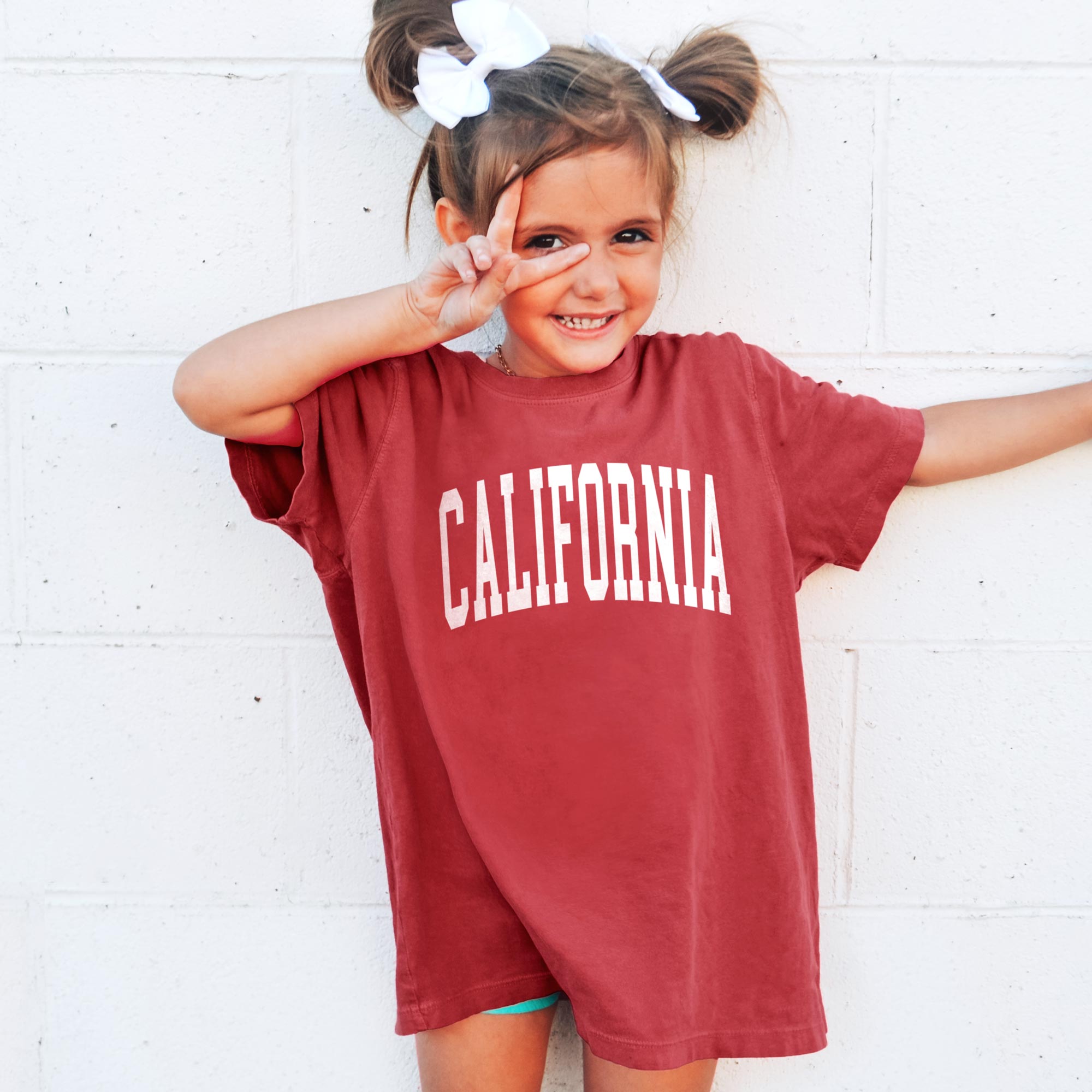 Young girl smiling and giving peace sign wearing red tee that says California.