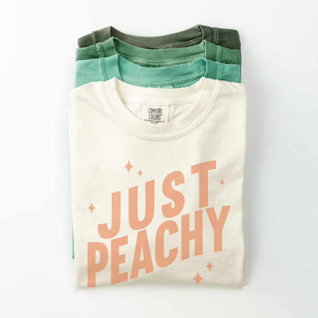 Stack of Comfort Color tees with the top one saying Just Peachy.